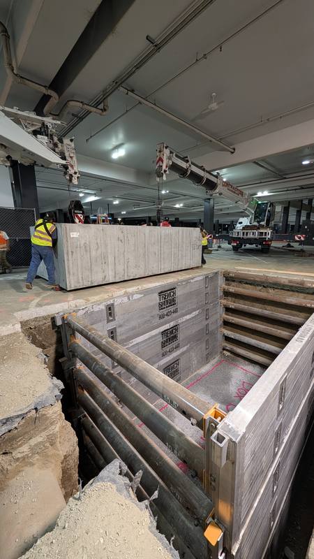Two cranes lifting a large precast concrete grease interceptor beside a shored excavation inside a parking structure
