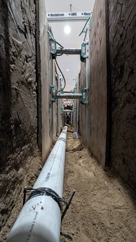 Looking down a deep, narrow shored trench with new white sewer pipe on the bottom